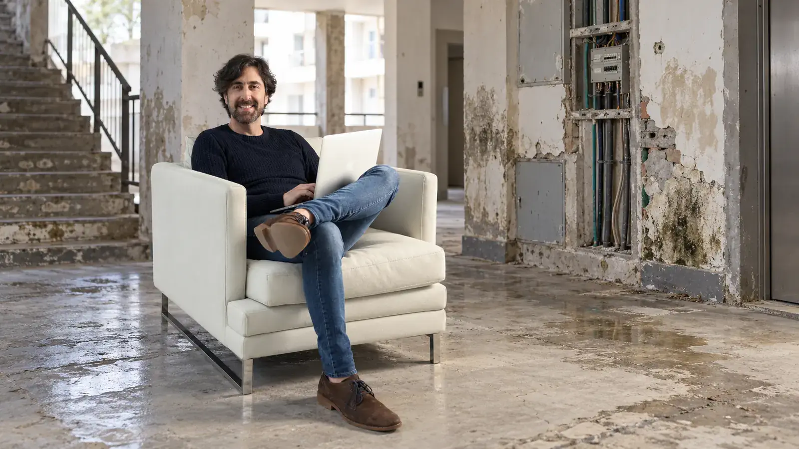 Elbio Stoler sentado con una computadora en una composición editorial con un edificio deteriorado de fondo