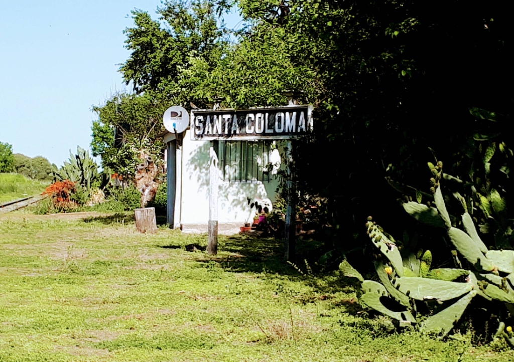 Fotografía del histórico cartel nomenclador de la estación Santa Coloma, rodeado de cactus y vegetación frondosa.